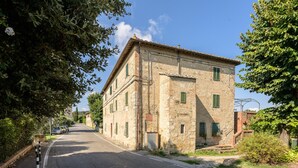 Ein zweistöckiges Steinhaus mit grünen Fensterläden, eine gepflasterte Straße davor und Bäume an den Seiten.
