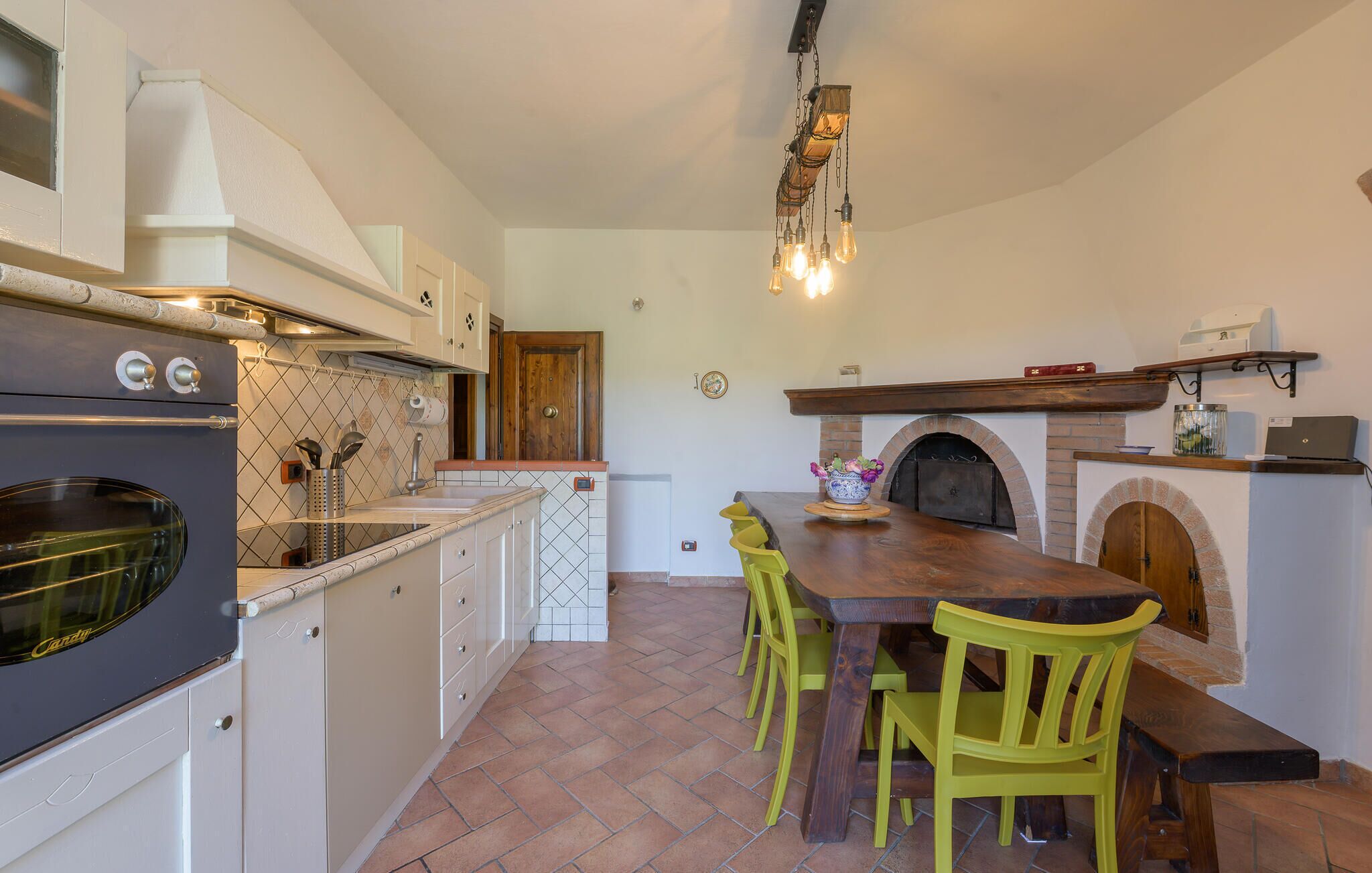 A kitchen with a large wooden table, two green chairs, a built-in oven, and a brick fireplace.