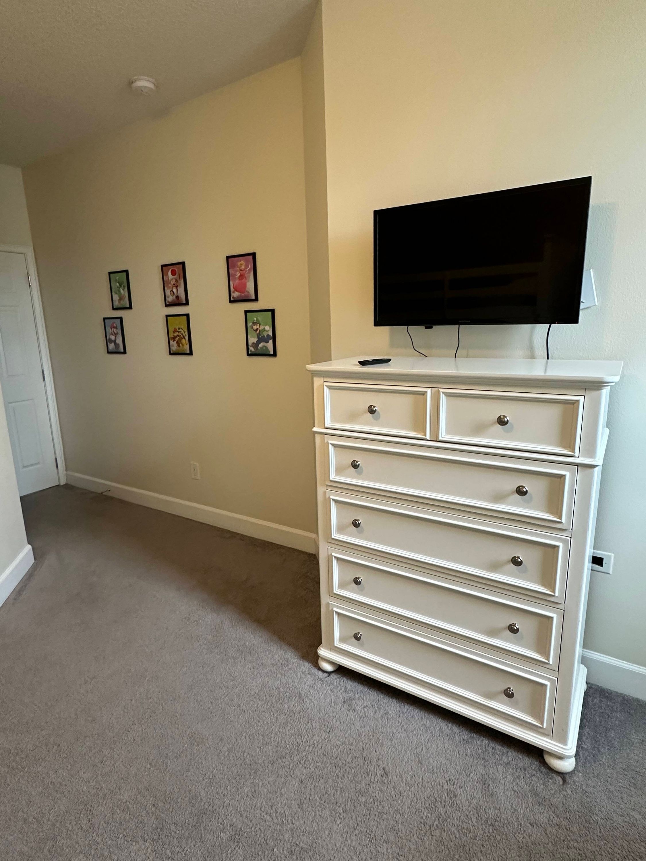 A room with a white dresser, a flat-screen TV mounted on the wall, and four framed pictures on the wall.