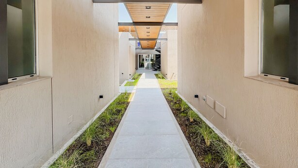 A modern corridor with a concrete pathway flanked by grass and plants, leading to a building entrance. - Casa Foresta (Salta)