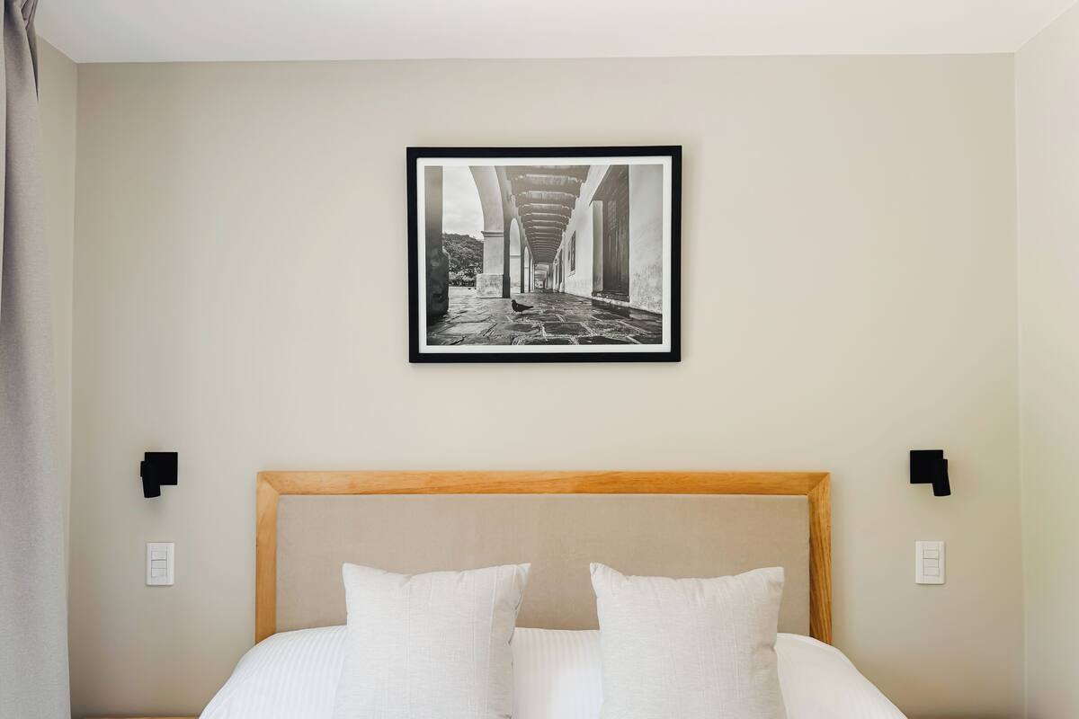 A bedroom with a bed, two pillows, a wooden headboard, and a framed photograph on the wall.