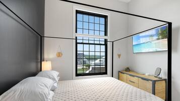 Un dormitorio con una cama grande, un escritorio con computadora, un televisor y una ventana con vista a una casa y árboles.