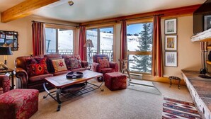 A cozy living room with a large window overlooking a snowy landscape, a fireplace, and a red sofa with patterned cushions. - Cozy 2BR Condo | Stunning Slope Views (Crested Butte)