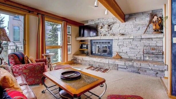 A cozy living room with a stone fireplace, a television, and a large window with curtains. - Cozy 2BR Condo | Stunning Slope Views (Crested Butte)