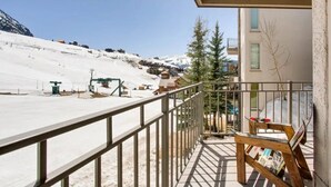 A balcony with a view of a snowy mountain slope, a wooden chair, and a small table. - Cozy 2BR Condo | Stunning Slope Views (Crested Butte)