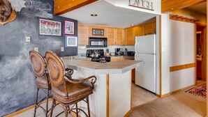 A kitchen with wooden cabinets, a white refrigerator, and a bar area with two stools. - Cozy 2BR Condo | Stunning Slope Views (Crested Butte)