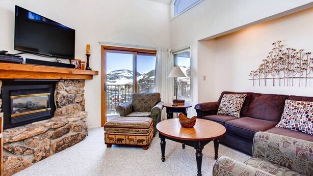 A living room with a stone fireplace, a television, a sofa, and a view of mountains through a balcony. - Rustic Mountain Retreat with Stunning Views (Crested Butte)