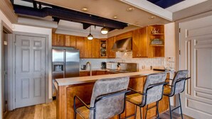 A modern kitchen with wooden cabinets, a stainless steel refrigerator, a built-in island with bar stools, and pendant lighting. - Modern 3BR Condo | Stunning Mountain Views (Crested Butte)