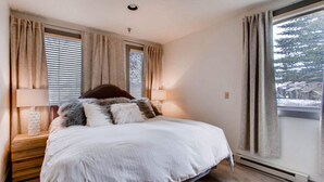 A bedroom with a large bed, two windows with curtains, a bedside table with a lamp, and a view of trees outside. - Modern 3BR Condo | Stunning Mountain Views (Crested Butte)