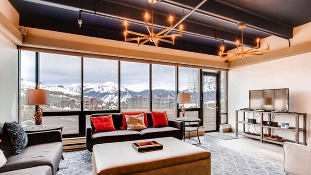 A modern living room with a large window, a sofa, a coffee table, and a TV stand. - Modern 3BR Condo | Stunning Mountain Views (Crested Butte)