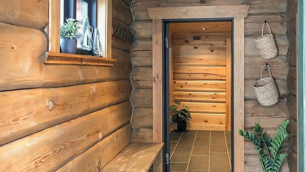 Interior de una cabaña de madera con un banco, una puerta que da a otra habitación y una planta en maceta.