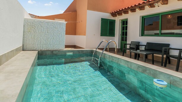 Una piscina amb un solàrium d'azulejos, una paret blanca amb un dibuix geomètric i un edifici amb teulada vermella i persianes verdes.