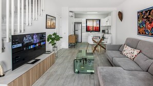 Una sala de estar moderna con una televisión de pantalla plana, un sofá gris, una mesa de centro de vidrio y una cocina al fondo.