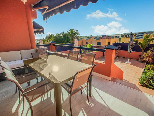 An outdoor patio with a table and chairs, a sofa, and a railing overlooking a residential area with houses and greenery.