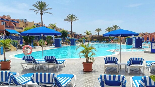 Un área de piscina con sillones de descanso y sombrillas, rodeada de palmeras y edificios.