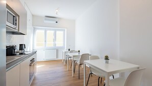 Eine moderne Küche mit weißen Schränken, einem Einbauofen und einer Mikrowelle. Ein Essbereich mit weißen Tischen und Stühlen, und ein Fenster sorgt für natürliches Licht.
