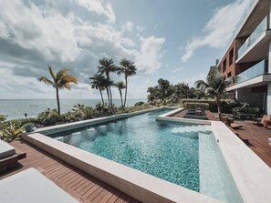 A pool with clear water, surrounded by palm trees and a view of the ocean. - BT Homes - Elegant Beachfront Apartment (Tulum)