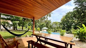 An outdoor patio with a wooden table and benches, a hammock, and a pool in the background.