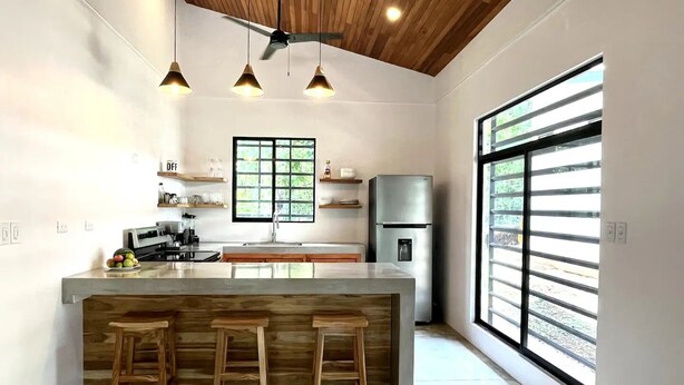A modern kitchen with a central island, wooden stools, a refrigerator, and a large window with blinds.