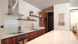 A modern kitchen with wooden cabinets, a white countertop, and a built-in oven. There are shelves with kitchenware and a hanging macrame decoration. - Luxury House in the Jungle with Pool by BT Homes (Tulum)