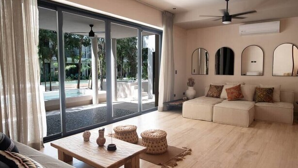 A spacious living room with a sofa, a coffee table, and a view of a pool and greenery outside. - Luxury House in the Jungle with Pool by BT Homes (Tulum)