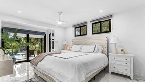A modern bedroom with a large bed, white nightstands, a ceiling fan, and a view of a swimming pool.