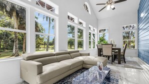A spacious living room with a beige sectional sofa, a glass coffee table, and a dining area with a table and chairs. Large windows provide views of the outdoors.