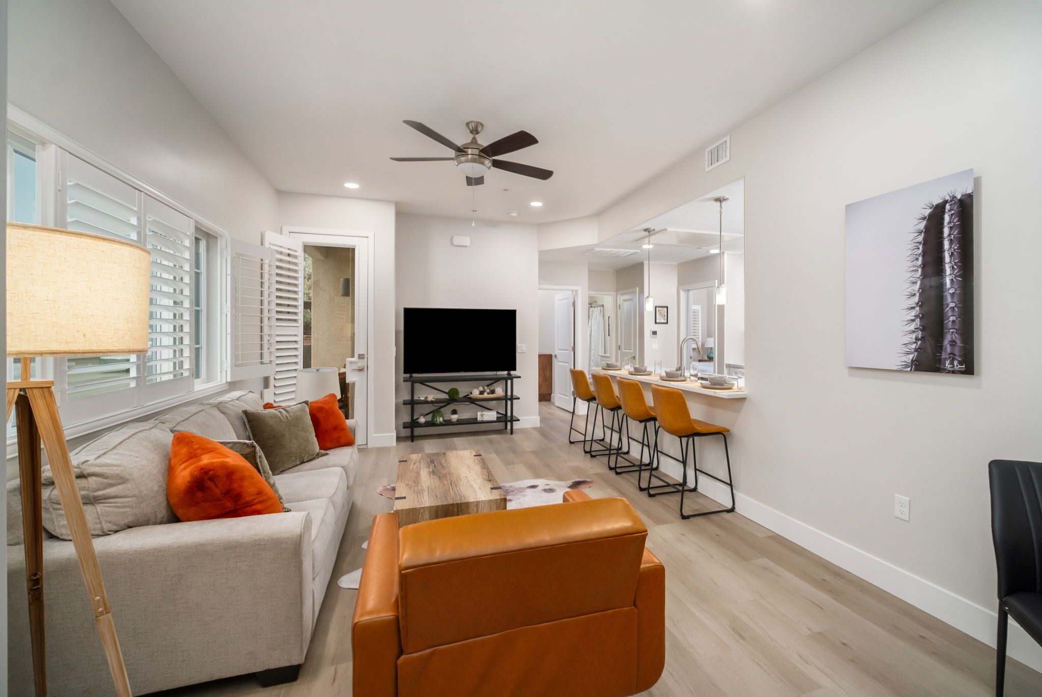 A modern living room with a sofa, coffee table, and orange armchair. There is a flat-screen TV, a ceiling fan, and a dining area with chairs.