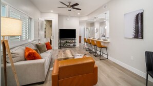 Ett modernt vardagsrum med en soffa, ett soffbord och en orange fåtölj. Här finns också en platt-TV, en takfläkt och en matplats med stolar.