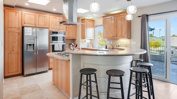 A modern kitchen with wooden cabinets, a stainless steel refrigerator, a built-in oven, and a large island with stools. - Beach Dream - Steps to the boardwalk & beach with AC, WiFi, deck, prime location! (San Diego)