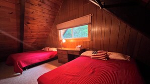 Una cabaña con dos camas, paredes de madera, una ventana con persianas y un escritorio con lámpara.