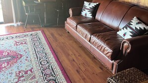 Ein gemütliches Zimmer mit einem braunen Ledersofa, einem gemusterten Teppich, Holzboden und einem Gemälde von Pferden an der Wand.