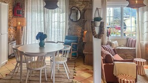 Un salon chaleureux avec un mur en pierre, une table ronde entourée de chaises, un fauteuil bleu, un canapé avec des coussins à rayures et une fenêtre donnant sur l’extérieur.