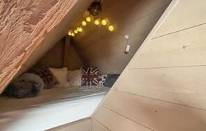 A cozy attic bedroom with a sloped ceiling, a bed with patterned pillows, and a string of warm lights.