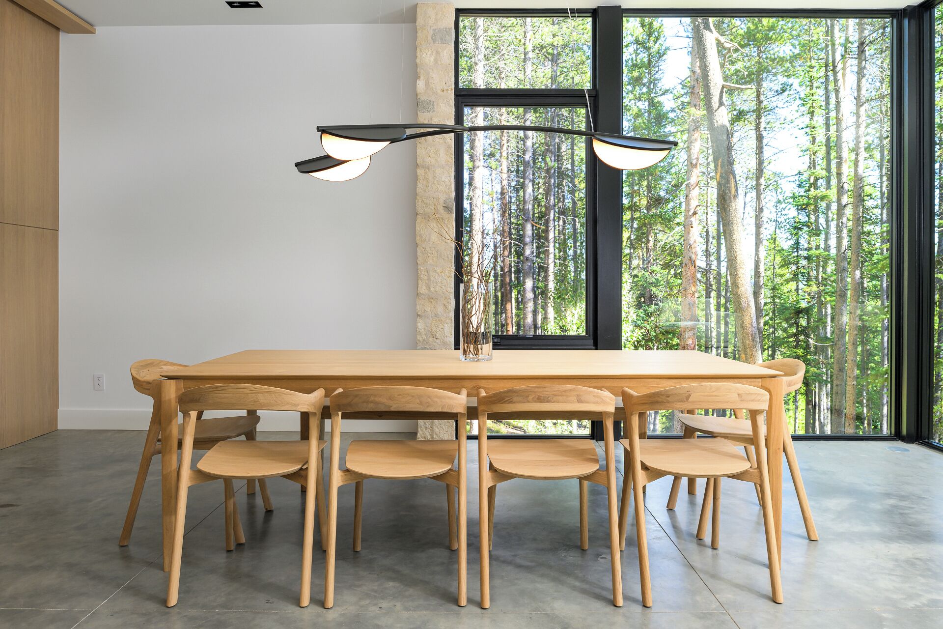 A dining area with a wooden table and chairs, a modern pendant light, and a large window with a forest view.