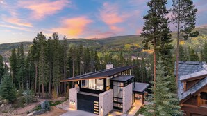 A modern two-story house with a large garage, surrounded by tall pine trees and a mountainous backdrop.