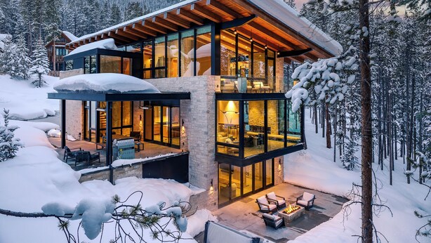 A modern, multi-level cabin with glass walls, a wooden roof, and a fireplace in the outdoor seating area.