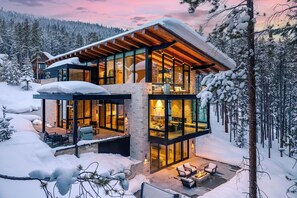A modern, multi-level cabin with glass walls, a wooden roof, and a fireplace in the outdoor seating area.