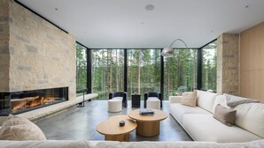 A modern living room with a stone fireplace, a large window offering a forest view, a white sofa, and two round wooden tables.