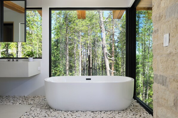 A modern bathroom with a freestanding bathtub, a white sink, and a large window offering a view of a forest.