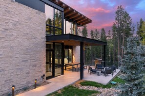 Une maison moderne avec une façade en pierre, de grandes fenêtres et un patio extérieur couvert, aménagé avec des sièges et un barbecue.