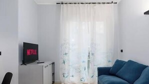 A modern living room with a blue sofa, a TV displaying the Netflix logo, and a white curtain with a leaf pattern. - [Center - Metro] Stylish and Modern Two-room apartment (Turin)