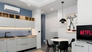 A modern kitchen with white cabinets, a black countertop, and a built-in oven. There is a dining area with a white table and black chairs. A black TV is mounted on the wall, and a framed artwork is visible. - [Center - Metro] Stylish and Modern Two-room apartment (Turin)