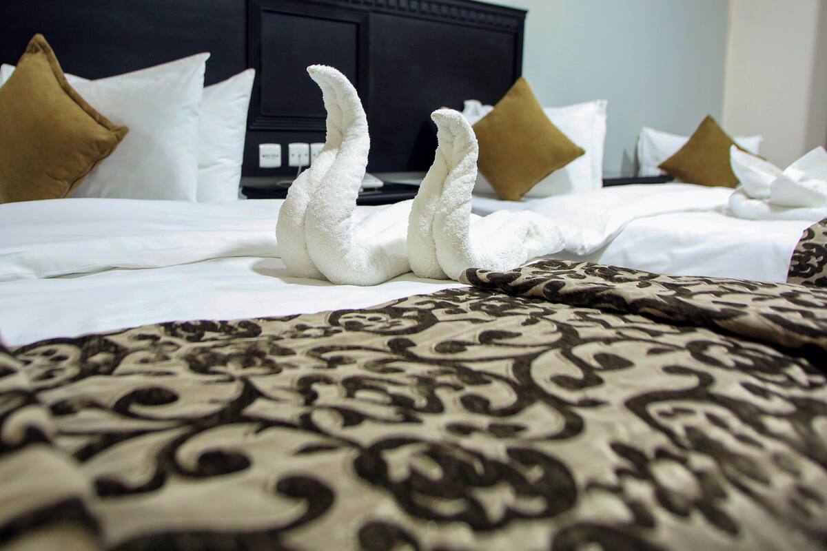 A hotel room with a neatly made bed, a patterned throw blanket, and a dark headboard.