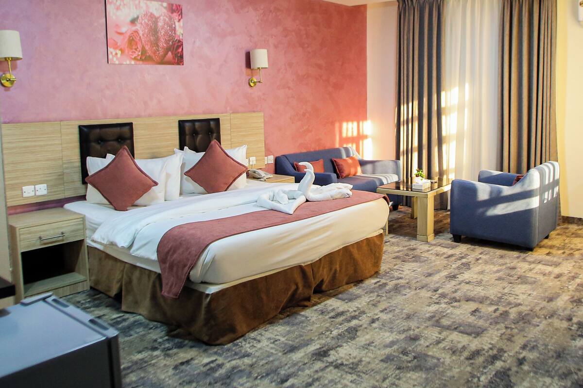 A hotel room with a large bed, a sofa, a desk, and a chair. There is a painting on the wall and a lamp on the nightstand.