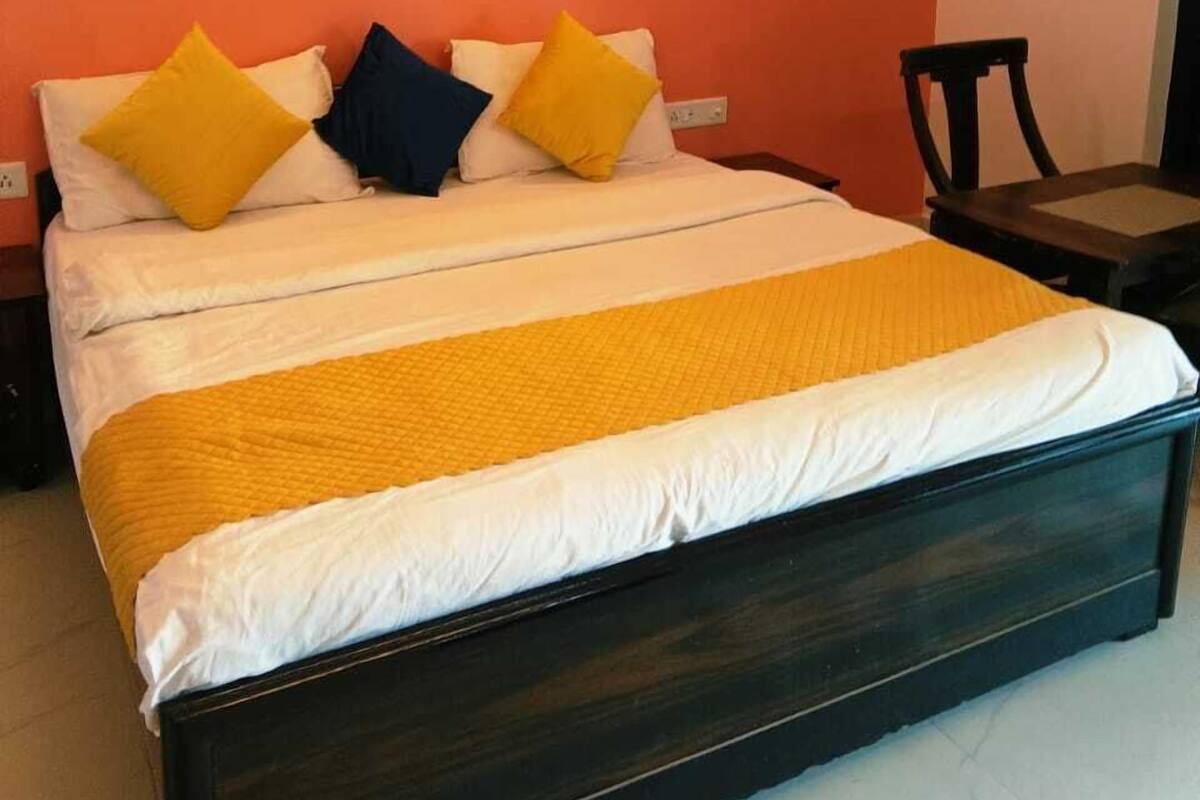 Una cama con estructura de madera oscura, ropa de cama blanca y amarilla, y tres almohadas en amarillo, azul y blanco.