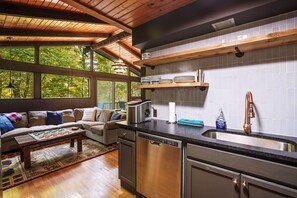 A modern kitchen with a dishwasher, a sink, and a coffee maker. There is a wooden ceiling and a window overlooking a garden.