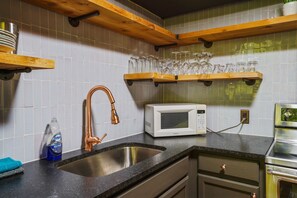A kitchen with a stainless steel sink, a white microwave, and wooden shelves holding glassware.