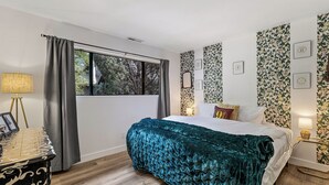 Un dormitorio con una cama grande, una ventana con cortinas, una mesita de noche con lámpara y papel tapiz con motivos florales.