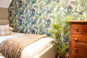 Une chambre à coucher avec un lit, une commode en bois et un mur recouvert d’un papier peint aux motifs tropicaux.